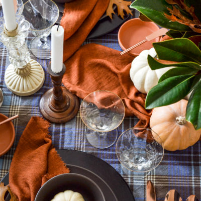 Overhead view of candles in wood and glass candlesticks and gold rimmed wine glasses, with terracotta colored napkins.
