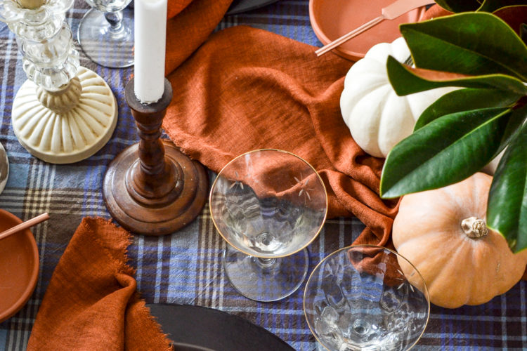 Overhead view of candles in wood and glass candlesticks and gold rimmed wine glasses, with terracotta colored napkins.