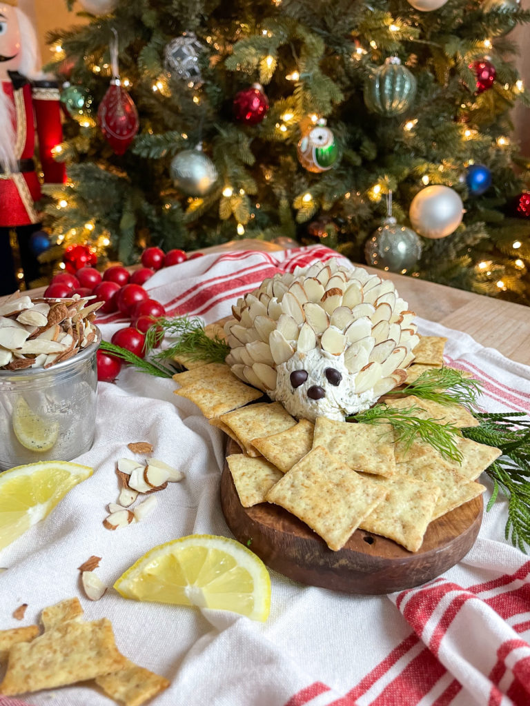 A hedgehog cheese ball surrounded by crackers served on a wood board in front of a Christmas tree