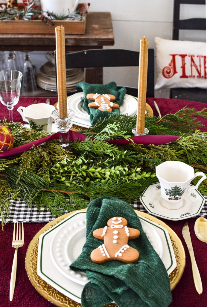 Christmas tablescapes with a table dressed in cranberry, gold and green.  A gingerbread man cookie sits on top of each pacesetting with fresh greens and gold candles down the centre.