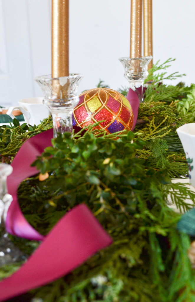 Fresh greens with a cranberry satin ribbon runs down the centre of a table.  Two gold taper candles are at one end and a colourful glass Christmas ornament in the middle.