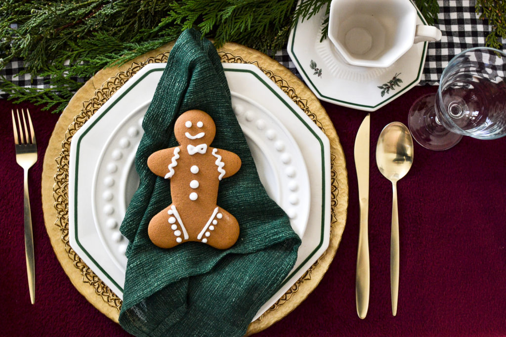 An over head view of a place setting with a gold charge under white plates.  A green napkin sits under a gingerbread man cookie.  Gold flatware at each side of the plates.