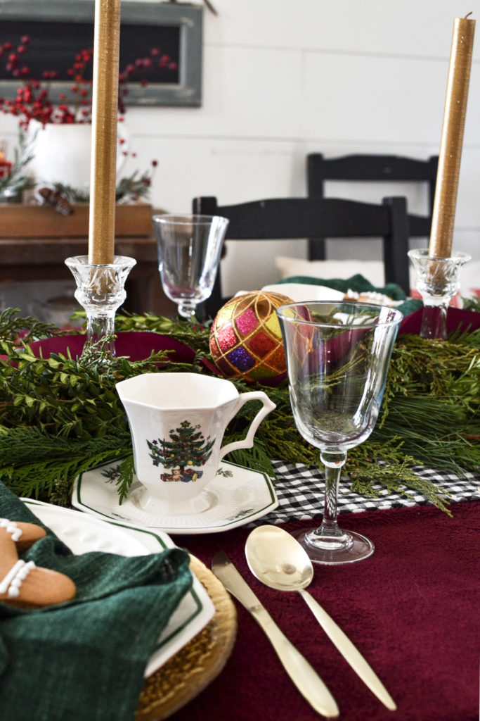 A bell shaped wine glass, a Christmas teacup with a Christmas tree on it, a black and white check table runner on top of a cranberry coloured tablecloth and fresh greens.