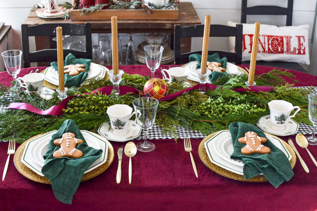 Christmas tablescapes with a cranberry tablecloth, white plates on gold chargers, topped with a green napkin and gingerbread man cookie