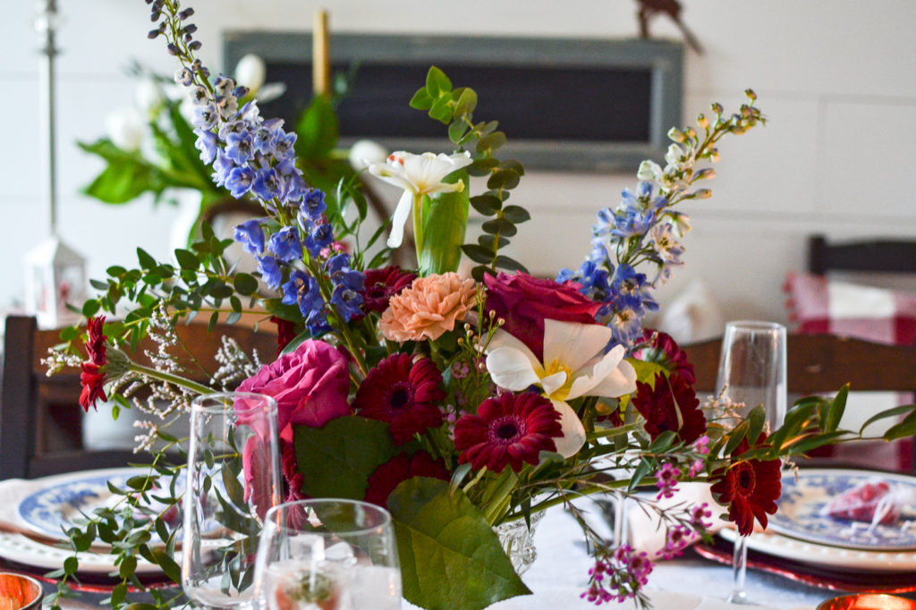 A brightly coloured floral centerpiece with roses, carnations and delphinium.