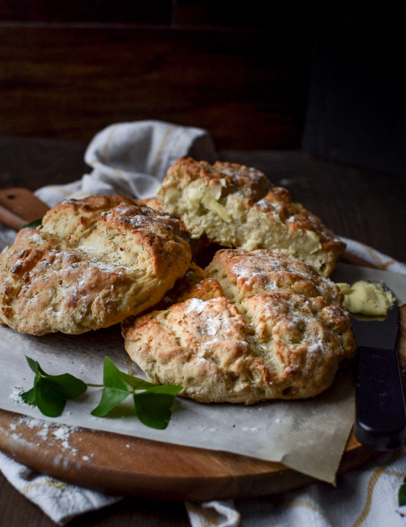 3 loaves of Irish Boxty Bread on a round cutting board. One is sliced open with butter spread on it.