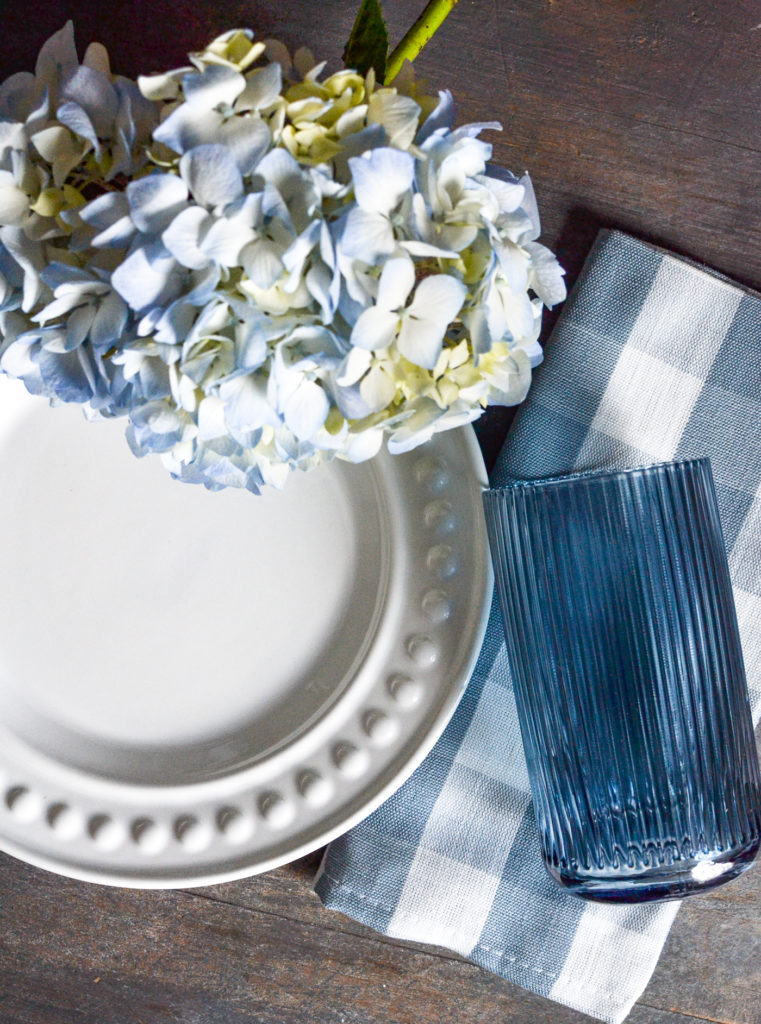 Overhead view of a white dinner plate, blue hydrangrea, blue and white check napkin and a blue glass.