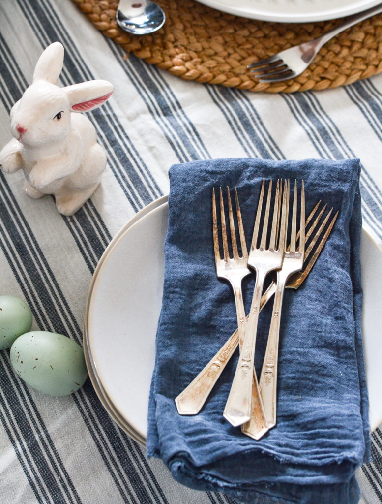 Blue striped tablecloth, a stack of plates with blue napkins and silver forks and a little white ceramic bunny. rabbit