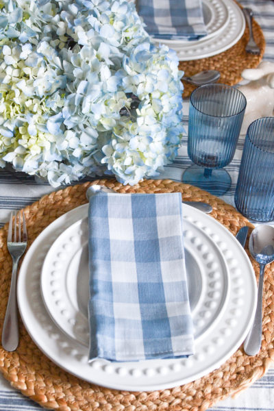 A soft blue and white checked napkin on top of white plates, with blue wine and high ball glasses on the side.