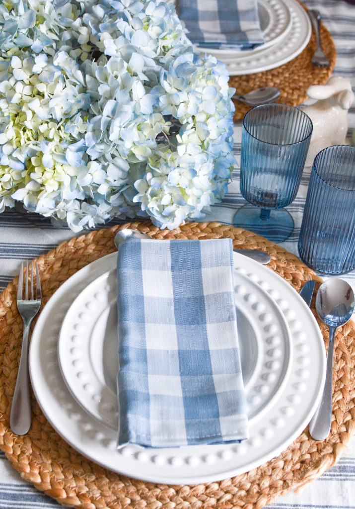 A soft blue and white checked napkin on top of white plates, with blue wine and high ball glasses on the side.