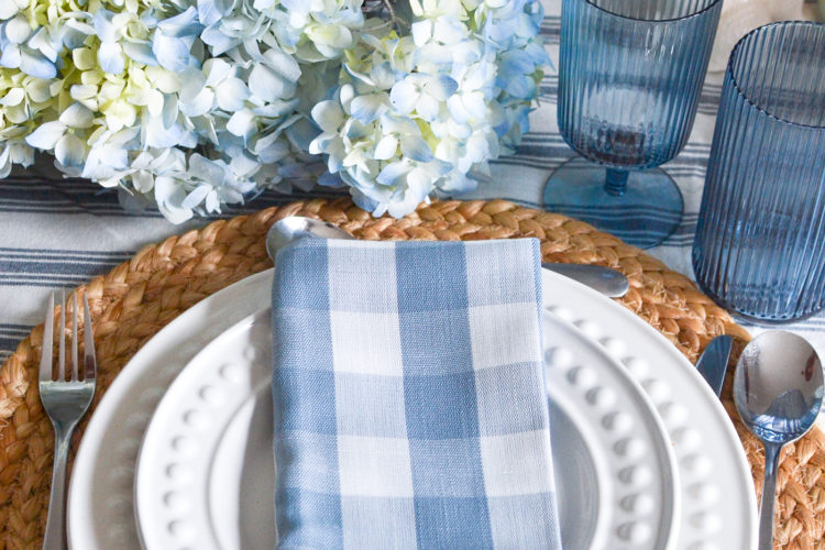 A soft blue and white checked napkin on top of white plates, with blue wine and high ball glasses on the side.