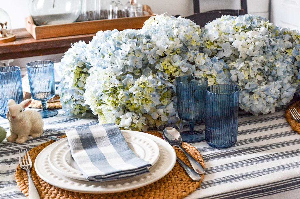 A blue and white tablescape with blue hydrangea centrepiece, white plates and blue and white checked napkins, and blue glassware.