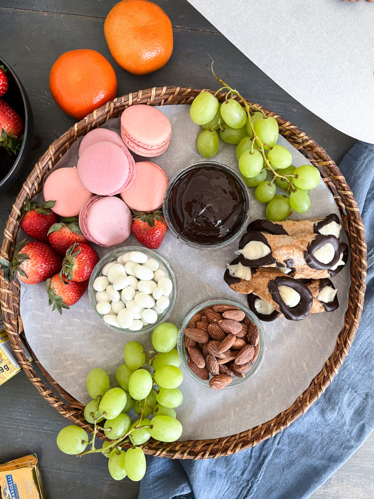 Begin filling in the dessert board with grapes, strawberries. Fill the bowls with chocolate dip, nuts and yogurt covered raisins