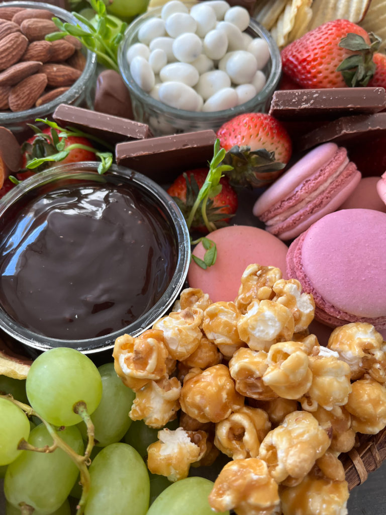 Close up of a dessert board with caramel corn, pink macarons, grapes, strawberries and chocolate dip.
