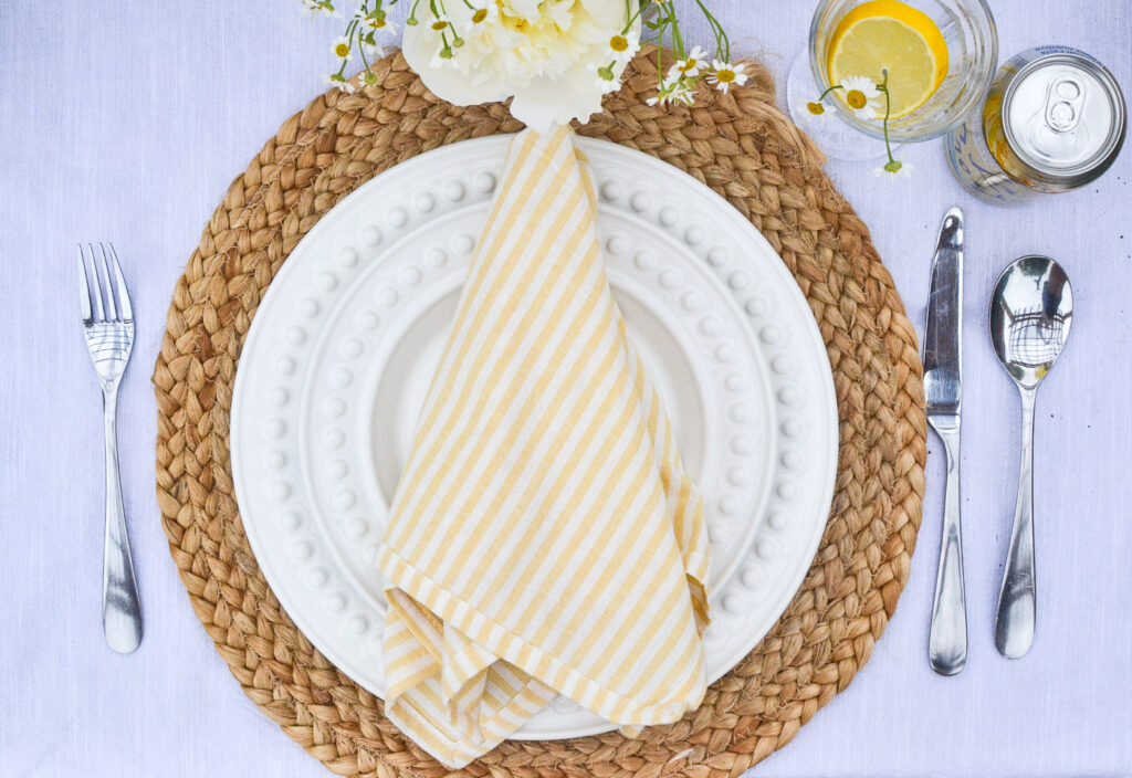 White dinner plate on a round rattan place mat, with a yellow and white striped napkin on top of the plate.