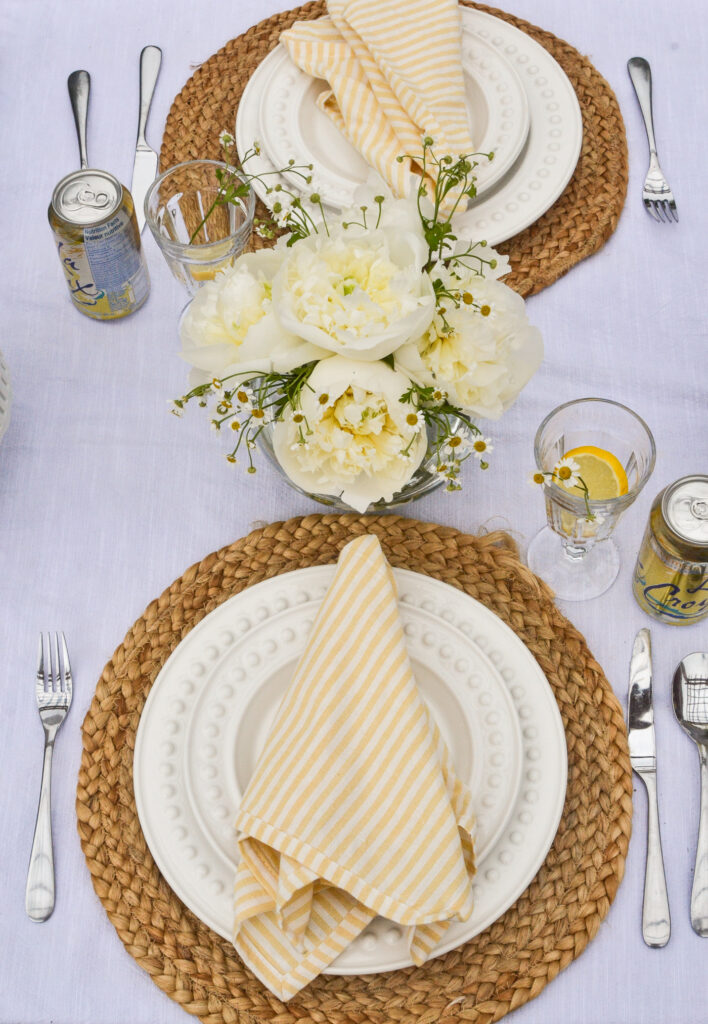 Two place settings of white dinner plates and yellow and white striped napkins, with a centrepiece of white peonies and chamomile flowers.