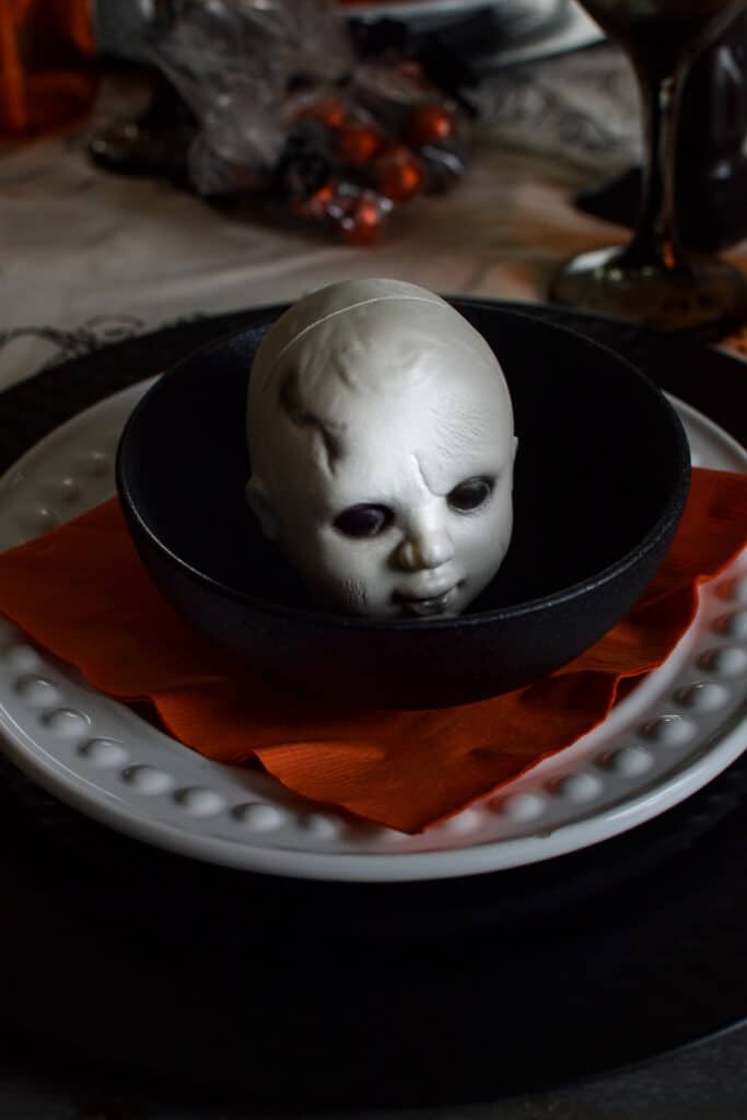 Black bowl with a spooky doll head in it on a table set for Halloween
