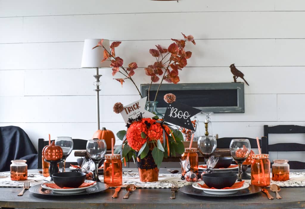 A dining table set with an orange and black theme including black bowls, orange napkins, orange ball jar glasses and orange hydrangeas
