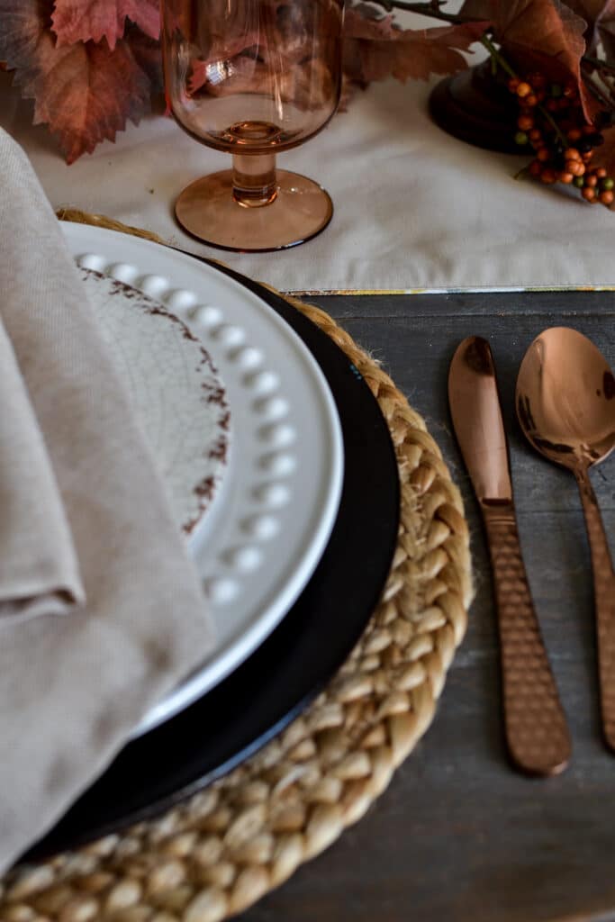 A place setting on a Thanksgiving table with a straw placemat, wood charger plate and white dinner plate.