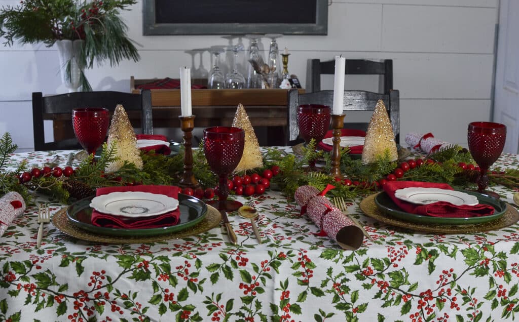 A Christmas tablescape with a holy tablecloth, red goblets and green stoneware dinner plates