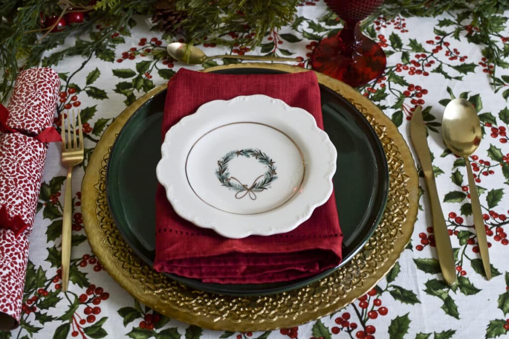 A place setting on a cottage style Christmas tablescape.  Gold charger with a green stoneware plate, red napkin and a holy wreath appetizer plate on top