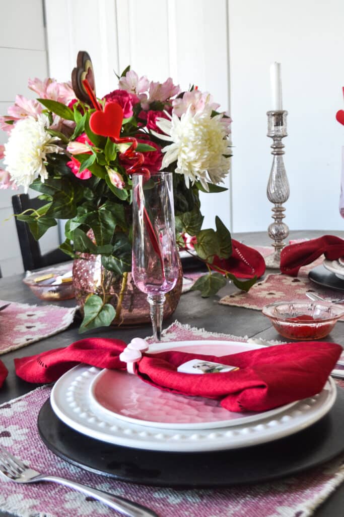 A Valentines Day place setting with a wood charger white dinner plate, pink salad plate and a red napkin topped with a miniature chocolate bar.