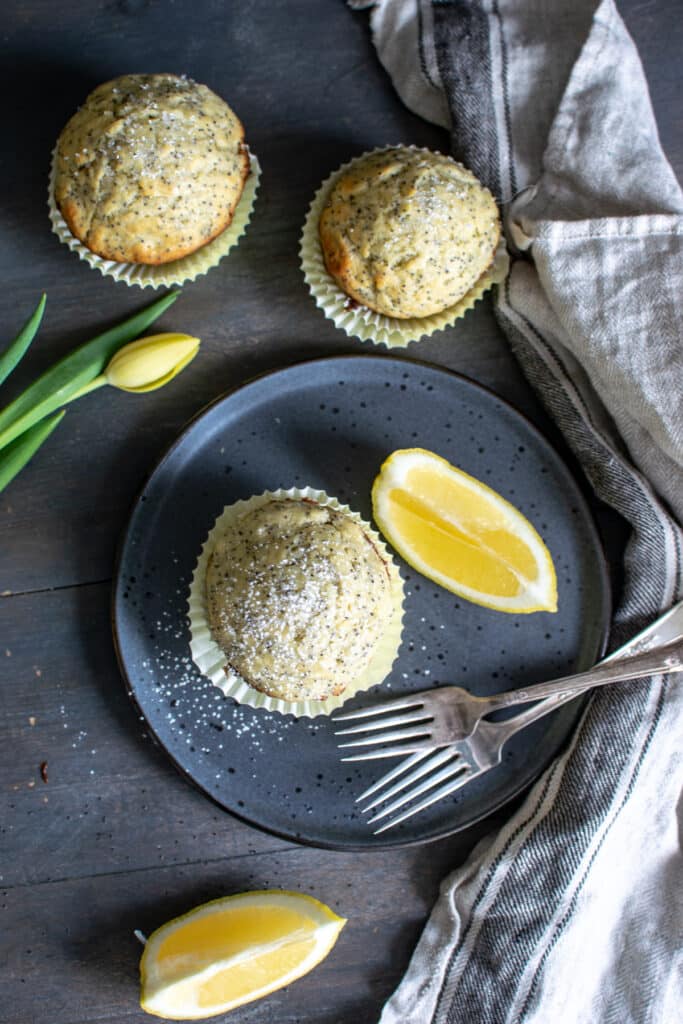 An over head view of lemon muffin with poppy seeds, with slices of lemons and 2 forks on a blue plate.