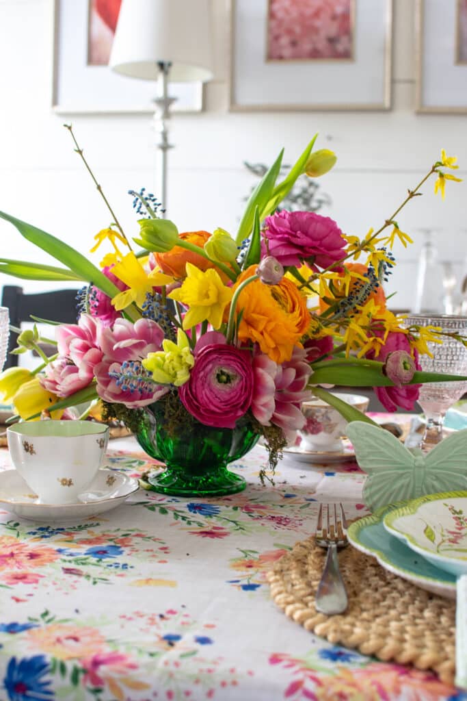 A vibrant floral centrepiece with orange ranunculus, yellow daffodils and pink tulips in the centre of a table set with spring dishes.