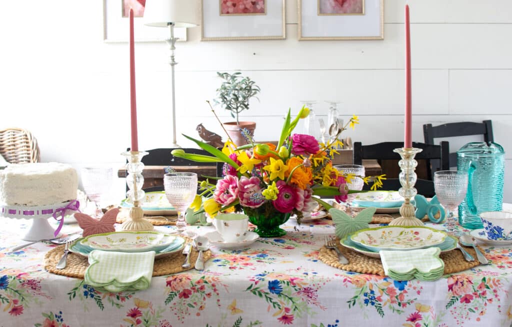 A brightly coloured floral table cloth covers a dining table set with floral themed plates and a bright coloured centrepiece of spring flowers.