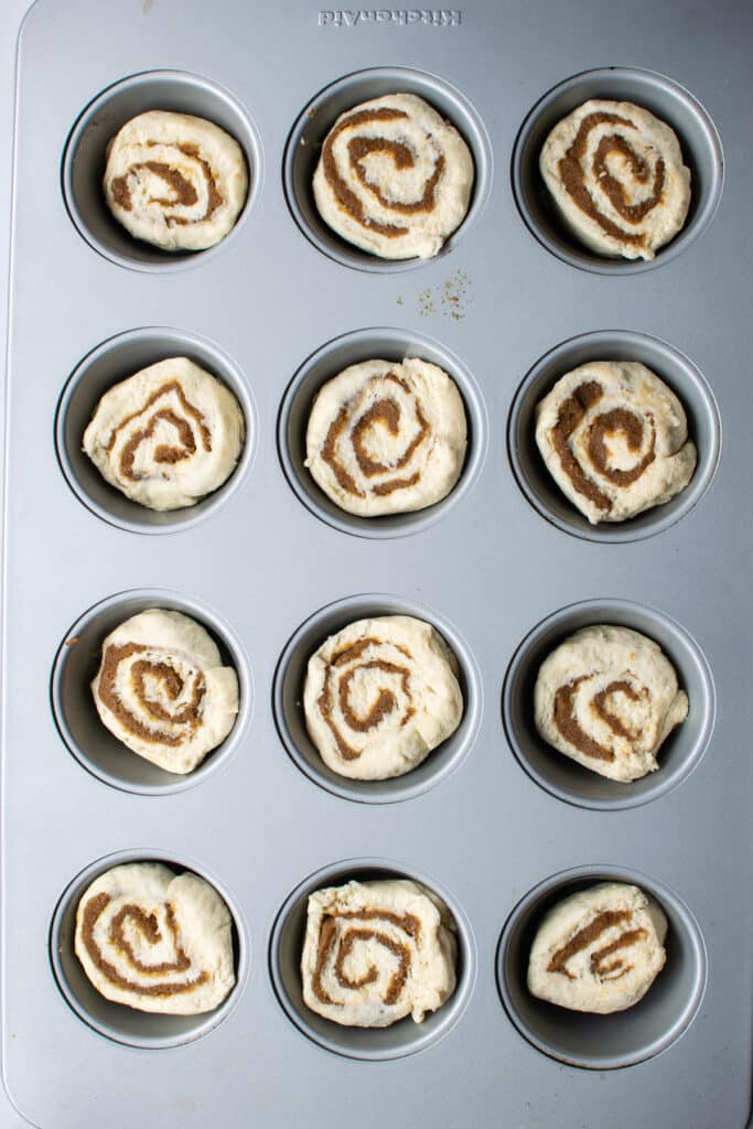 An overhead view of uncooked cinnamons cut and placed into 12 muffin tins ready to be baked.