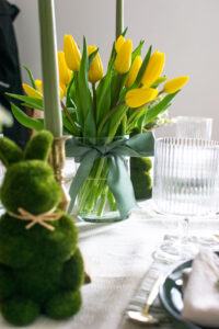 2 moss covered Easter bunnies with a vase of yellow tulips in the middle on a green tablescape.