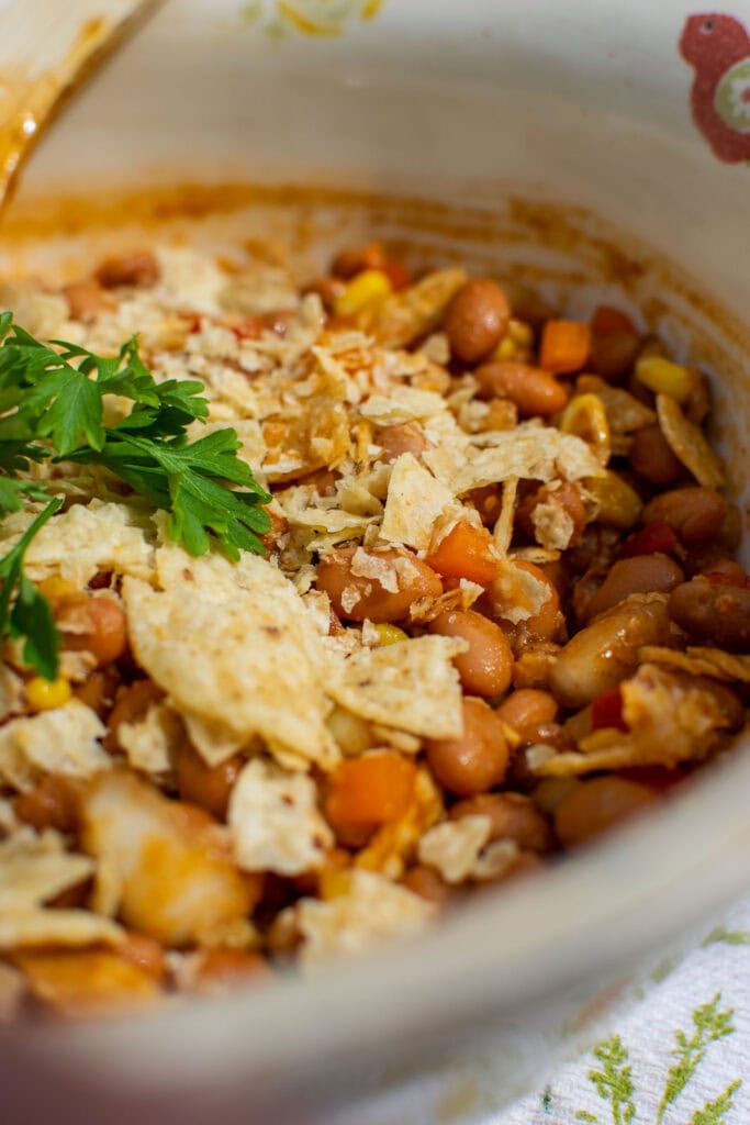 A bowl of cold baked bean salad topped with crushed tortilla chips and a sprig of parsley.