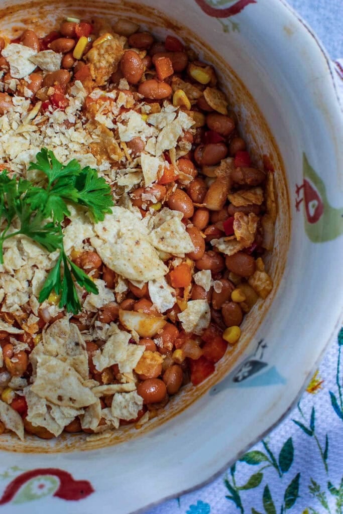 A large bowl filled with baked bean salad that is garnished with crushed tortilla chips and fresh parsley.