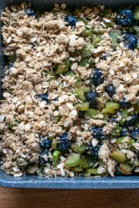 Crumble topping being sprinkled over top of blueberry rhubarb filling in a baking pan.