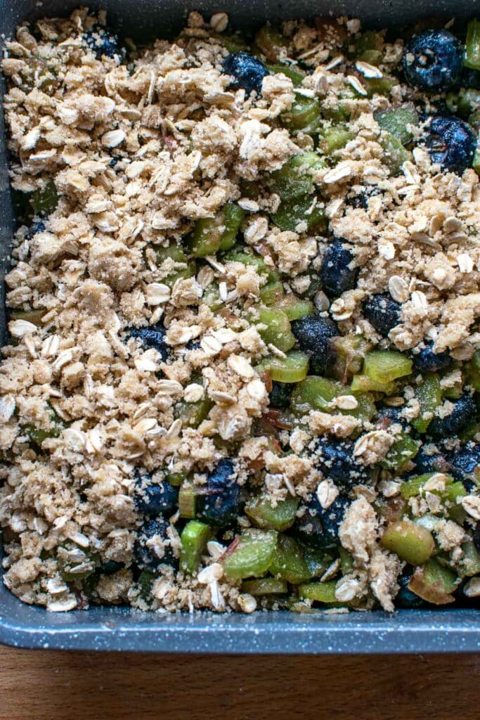Crumble topping being sprinkled over top of blueberry rhubarb filling in a baking pan.
