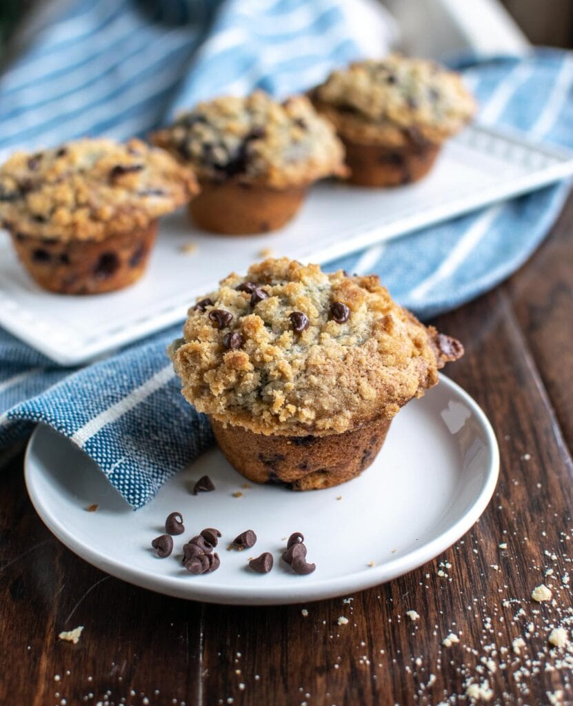 A blueberry chocolate chip muffin on a white plate with 3 more muffins in the background.