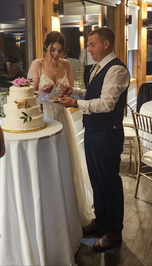 Bride and groom cutting wedding cake decorated with pink peonies and little golden retriever statues.