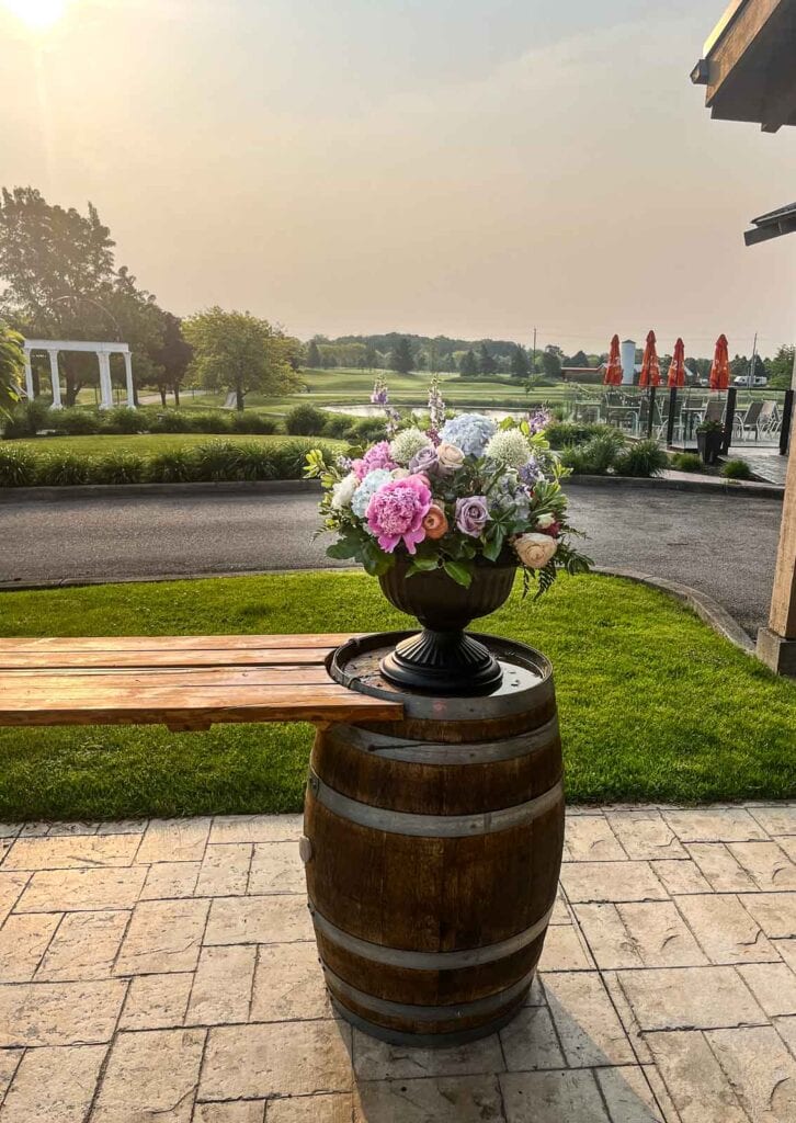 A large flower arrangement of peonies, hydrangeas and other flowers in pinks, purples and blues in an urn on top of wine barrels.