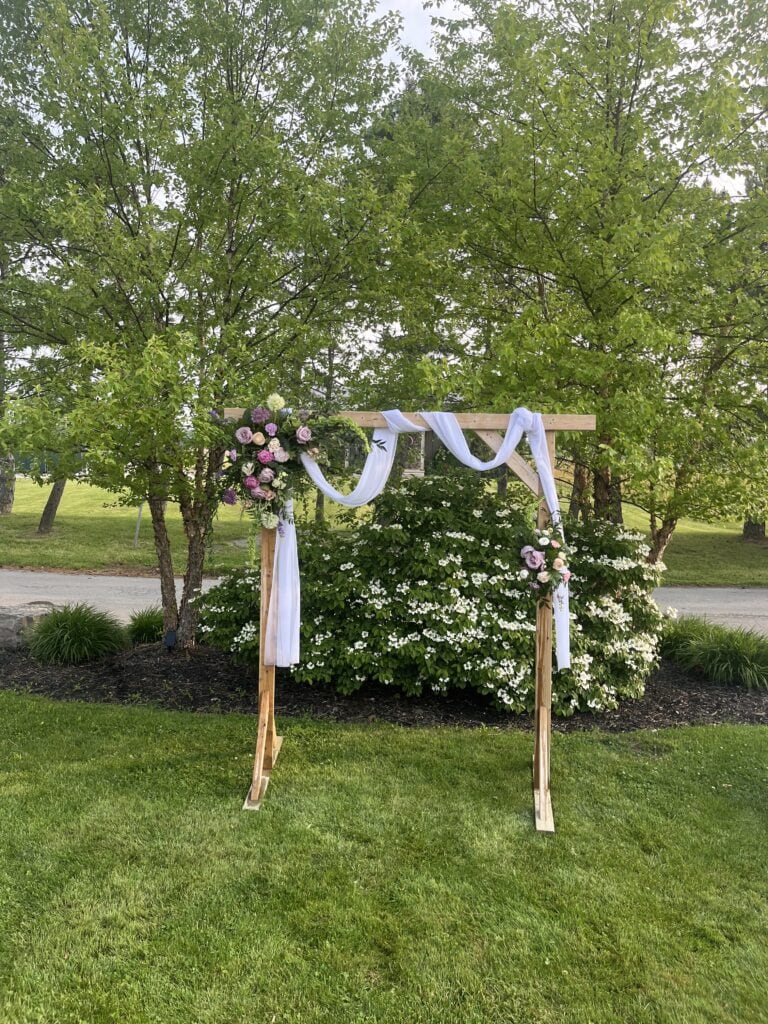 A wedding arbour decorated with pink, blue and purple flowers, and draped with a white sash.