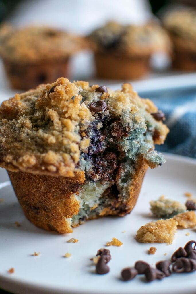 A blueberry chocolate chip muffin with a bit taken out of it on a white plate.