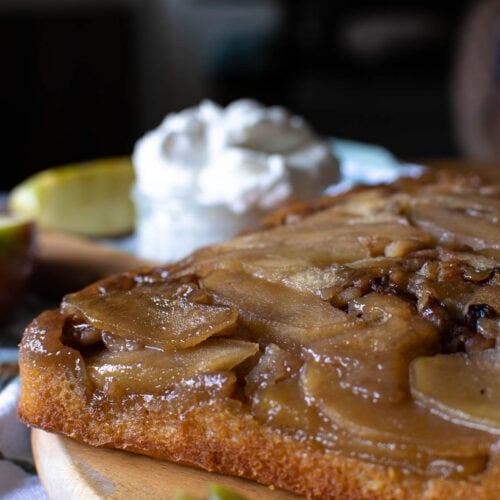 Maple Apple and walnut cake on a wooden platter with a bowl of whipped cream in the background.