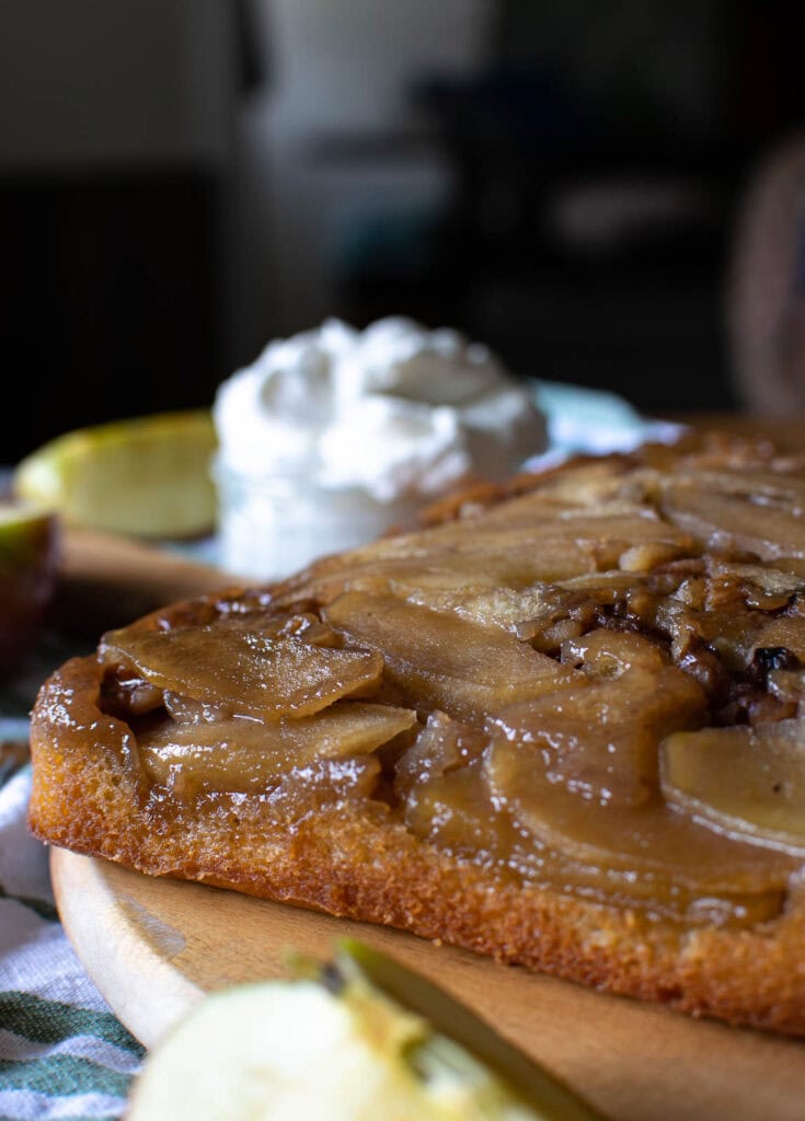 Maple Apple and walnut cake on a wooden platter with a bowl of whipped cream in the background.