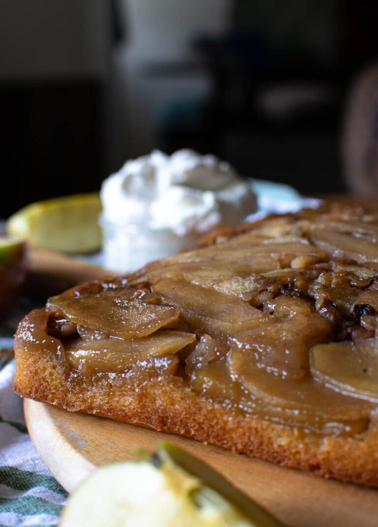 Maple Apple and walnut cake on a wooden platter with a bowl of whipped cream in the background.