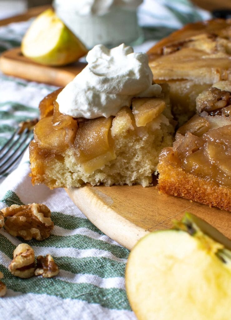 A slice of maple walnut cake pulled away from the full cake and topped with fresh whipped cream.