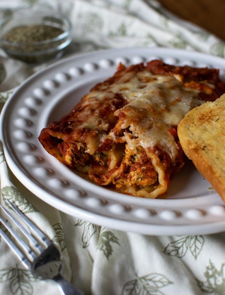 Cheesy chicken and spinach Manicotti on a white dinner plate with a side of garlic bread.
