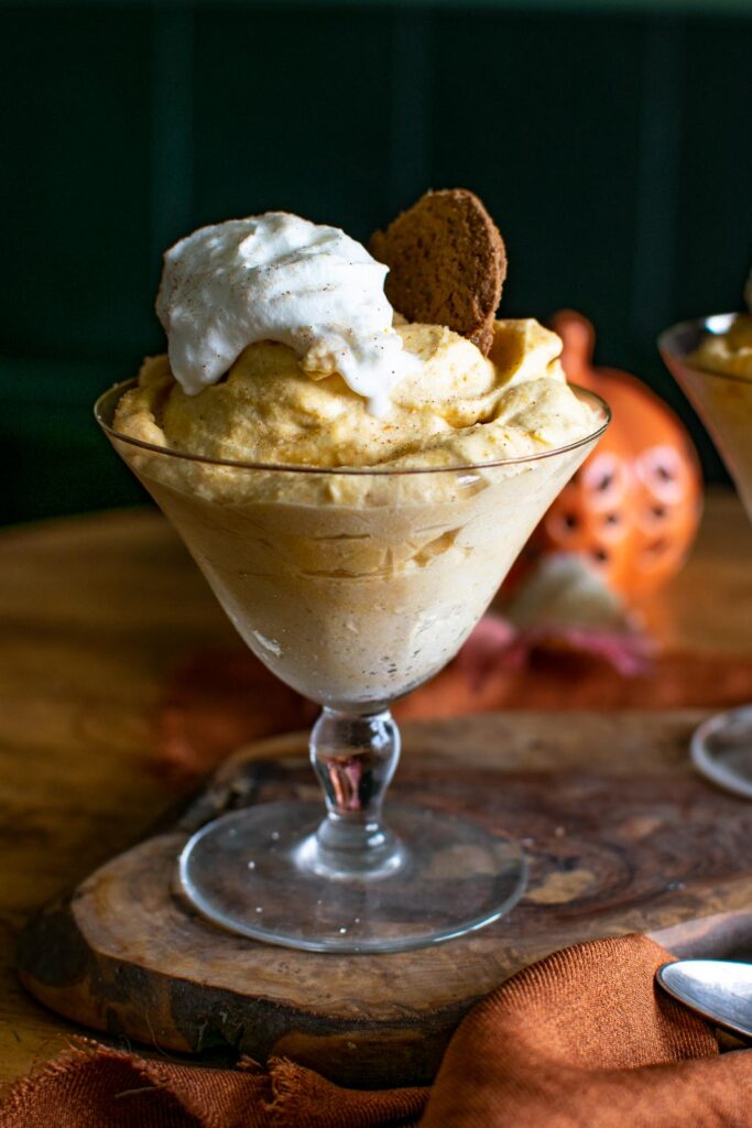 A footed dessert bowl filled with pumpkin mousse topped with whipped cream and a gingersnap cookie.