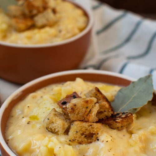 A close up of a bowl of cheesy cauliflower soup that is garnished with croutons and a bay leaf. Another bowl ca be seen in the background.