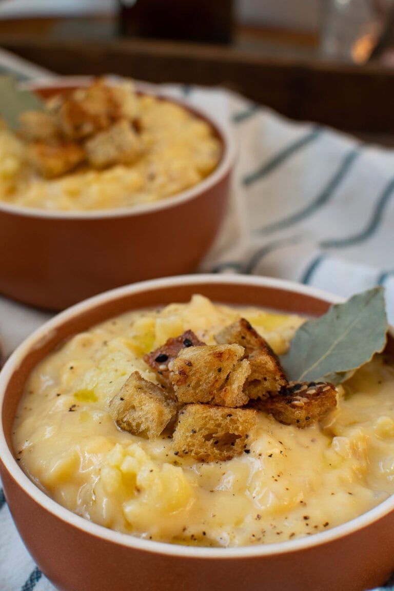 A close up of a bowl of cheesy cauliflower soup that is garnished with croutons and a bay leaf. Another bowl ca be seen in the background.