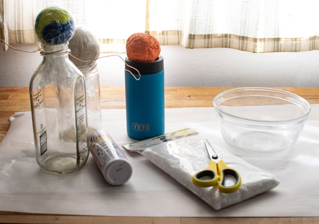 Round balls wrapped in plastic wrap sitting on tall jars to make ghosts using cheesecloth.