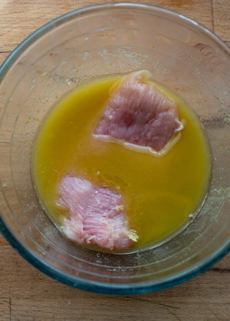A chicken breast fillet being dipped into seasoned melted butter mixture.